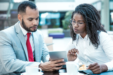 Focused mix raced colleagues watching content on tablet together during lunch. Business man and woman drinking coffee in cafe, using digital device, looking at screen. Lunch with colleague concept