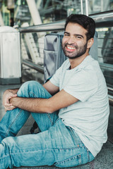 Smiling Hispanic man sitting on floor at airport. Cheerful young guy looking at camera while resting at terminal. Travel concept