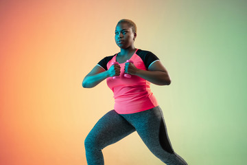 Young african-american plus size female model's training on gradient background in neon light. Doing workout exercises with the weights. Concept of sport, healthy lifestyle, body positive, equality.