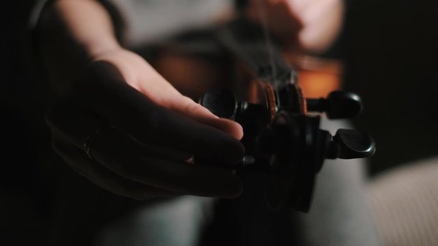 Young woman fiddler changing violin strings, loosing, twisting tuning peg at pegbox to unhook and pull the string out, moody low light scene at home