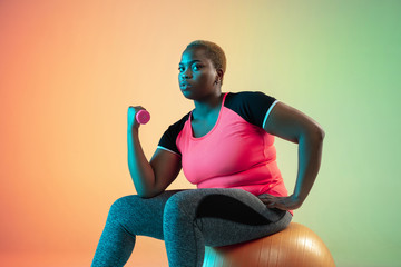 Young african-american plus size female model's training on gradient background in neon light. Doing workout exercises with fit ball. Concept of sport, healthy lifestyle, body positive, equality.