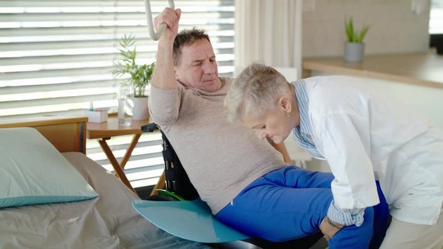 Healthcare worker helping paralysed senior patient into wheelchair in hospital.