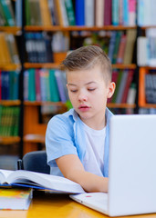 A boy sits in a library at a table, works on a laptop