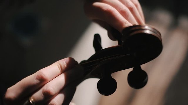 Young woman fiddler changing violin strings, tighten the string with the tuning peg at pegbox, moody low light scene at home