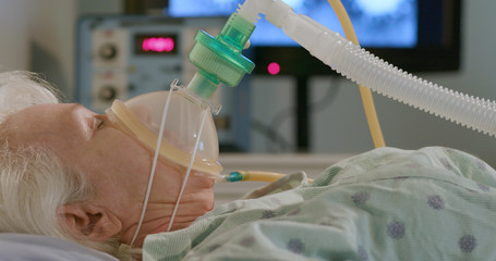 A hospitalized elderly woman attached to a device to help her breathe with an image of the coronavirus in the background.