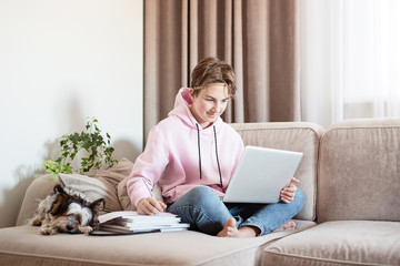 Distance learning online education. A schoolboy is studying at home with a laptop and doing school homework. Sitting on the sofa with textbooks. Nearby lies a dog - a pet