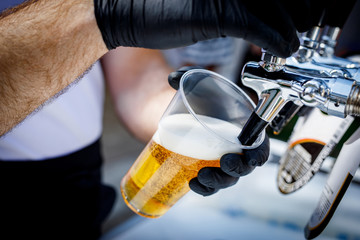 Bartender in black gloves pours beer into a disposable plastic glass