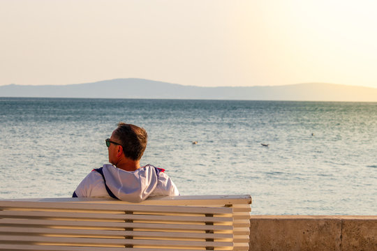 Older Man As Seen From Behind Sitting On A White Bench On Riviera Of Split, Croatia. Enjoying The Seascape View, Seagulls Flying Over The Sea