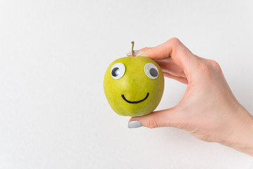 Female hand holding little Apple smile with Googly eyes. White background. Diet and weight loss...