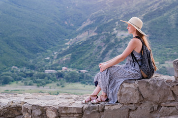 Naklejka premium Beautiful young woman in dress and hat sitting on background of mountain valley