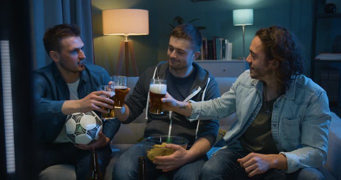 Three Caucasian guys sitting on sofa in dark living room with football ball and chips and watching football match on TV Best male friends spending evening with sport channel and doing cheers with beer