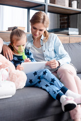 selective focus of asthmatic child using compressor inhaler and holding digital tablet near mother...