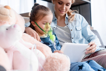 selective focus of asthmatic kid using respiratory mask and holding digital tablet near mother and soft toys