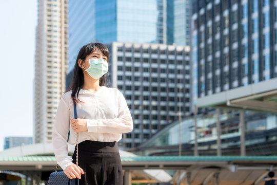 Young stress Asian businesswoman in shirt going to work in pollution city she wears protection mask prevent PM2.5 dust, smog, air pollution and COVID-19 with office building in Bangkok, Thailand.