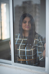 Portrait of young brunette Indian Bengali plus size woman in western wear looking outside in front of a glass window with water droplets/dewdrops on it. lifestyle and fashion.