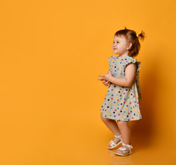Little ginger toddler girl with two ponytails, in gray polka dot dress, white sandals. Child smiling, posing on orange background