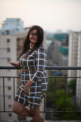Fototapeta na wymiar An young brunette Indian Bengali brunette plus size woman in western dress standing on rooftop/balcony in urban background while her hair is blowing in wind during sunset. lifestyle and fashion.