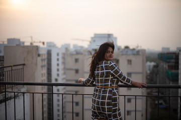An young brunette Indian Bengali brunette plus size woman in western dress standing on rooftop/balcony in urban background while her hair is blowing in wind during sunset. lifestyle and fashion.