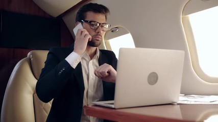 serious businessman typing on laptop and talking on smartphone in plane - Powered by Adobe