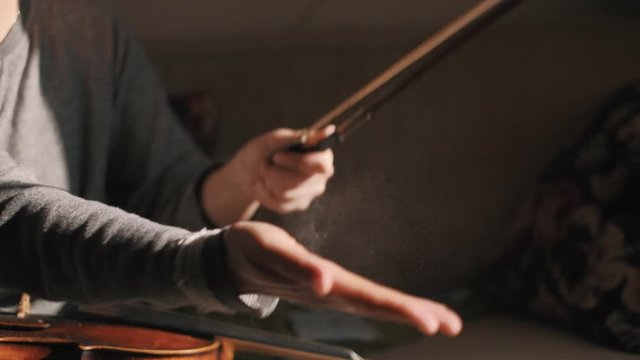 Young woman fiddler removing excess of rosin from the fiddlestick violin bow, closeup shot, dust specks particles floating in air