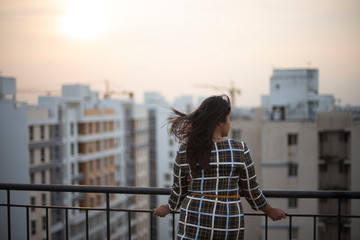 Fototapeta na wymiar Back side of an young brunette Indian Bengali brunette plus size woman in western dress standing on rooftop in urban background while her hair is blowing in wind during sunset. lifestyle and fashion.