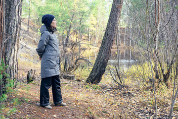 Girl in warm clothes with thoughtful look and hands behind her stands on the banks of taiga river in autumn forest.