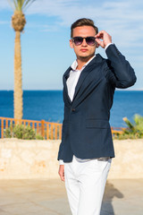 Young handsome man in a suit and white pants at the sea background and palm trees.