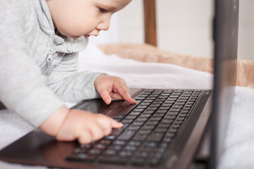  A little kid plays with a laptop, presses buttons, enjoys a new toy. A boy in a gray suit sits on a brown white canopy bed.
