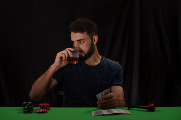 Portrait of young brunette Indian Kashmiri man in casual tee shirt playing cards on a casino poker table in black copy space studio background. lifestyle and fashion.