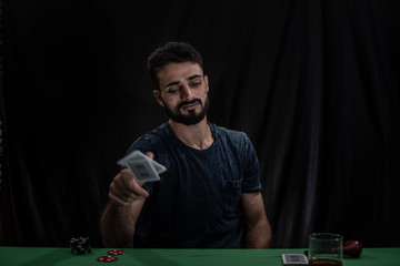 Portrait of young brunette Indian Kashmiri man in casual tee shirt playing cards on a casino poker table in black copy space studio background. lifestyle and fashion.