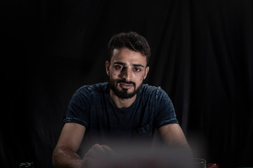 Portrait of young brunette Indian Kashmiri man in casual tee shirt playing cards on a casino poker table in black copy space studio background. lifestyle and fashion.