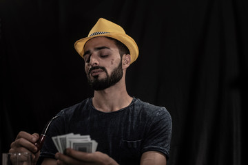 Portrait of young brunette Indian/European/Arabian/Kashmiri man in casual tee shirt and yellow hat playing cards on a casino poker table in black copy space studio background. lifestyle and fashion.