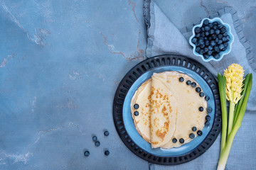 Crepes or pancakes with blueberries and butter on blue ceramic plate and yellow hyacinth flower on blue background. Top view or flat lay, copy space, horizontal