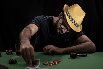 Portrait of young brunette Indian Kashmiri man in casual tee shirt and yellow hat playing cards on a casino poker table in black copy space studio background. lifestyle and fashion.
