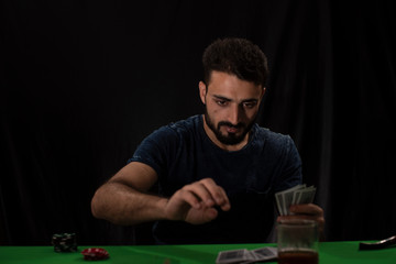 Portrait of young brunette Indian Kashmiri man in casual tee shirt playing cards on a casino poker table in black copy space studio background. lifestyle and fashion.