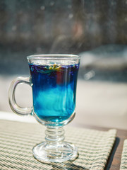 Amazing blue violet color butterfly pea flower blue tea in latte macchiato glass on window sill liten noon hard sunlight