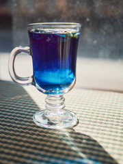 Amazing blue violet color butterfly pea flower blue tea in latte macchiato glass on window sill liten noon hard sunlight