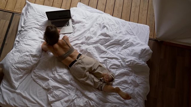 young beautiful girl with dark hair lies on a bed in a tent house. She works at a laptop and swings her legs.view from above.