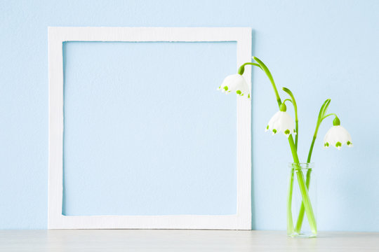 Fresh snowdrops in glass vase on table at light pastel blue wall. First messengers of spring. Empty place for inspirational, emotional, sentimental text, quote or sayings in white frame.