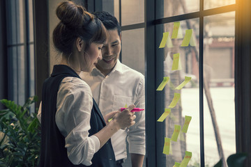Asian creative business female with businessman thinking with note and sticking at contact sheet on glass wall in office.