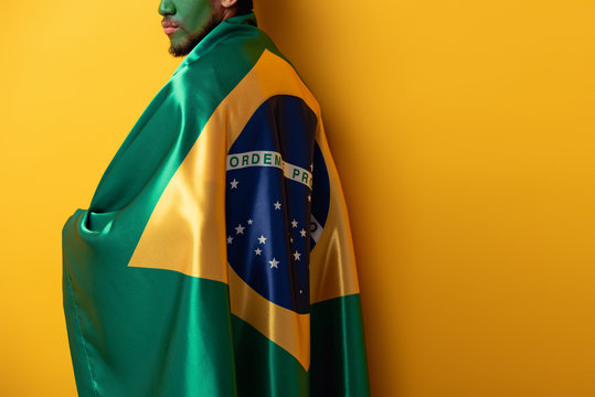 cropped view of football fan with painted face wrapped in brazilian flag on yellow