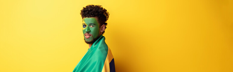 panoramic shot of african american football fan with painted face wrapped in brazilian flag on...