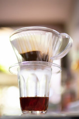 Drip brewing; clear glass of coffee mug and paper dripping bag with ground coffee in soft sunlight at home.