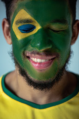 smiling african american football fan with closed eyes and face painted as brazilian flag isolated on grey
