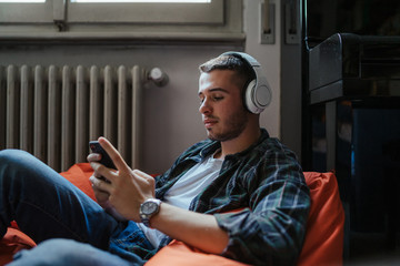 Young man listening to music with headphones and use your smartphone sitting on a pouf in her room...
