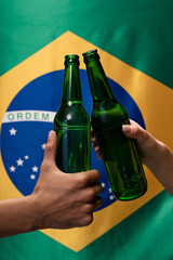 cropped view of couple of football fans clinking with bottles of beer with brazil flag behind