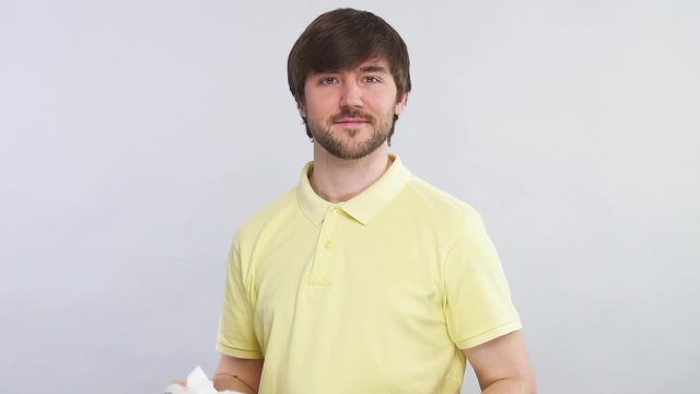 young bearded man in yellow shirt start to sneeze into napkin on grey background and showing thumbs up