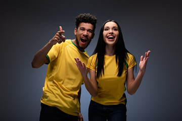 emotional multiethnic couple of football fans in yellow t-shirts gesturing on grey