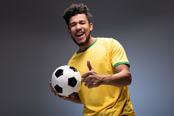 happy african american football fan in yellow t-shirt holding ball and showing thumb up on grey