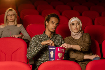 African american muslim man with his wife sitting in movie theater, watching movie, eating popcorn, smiling.